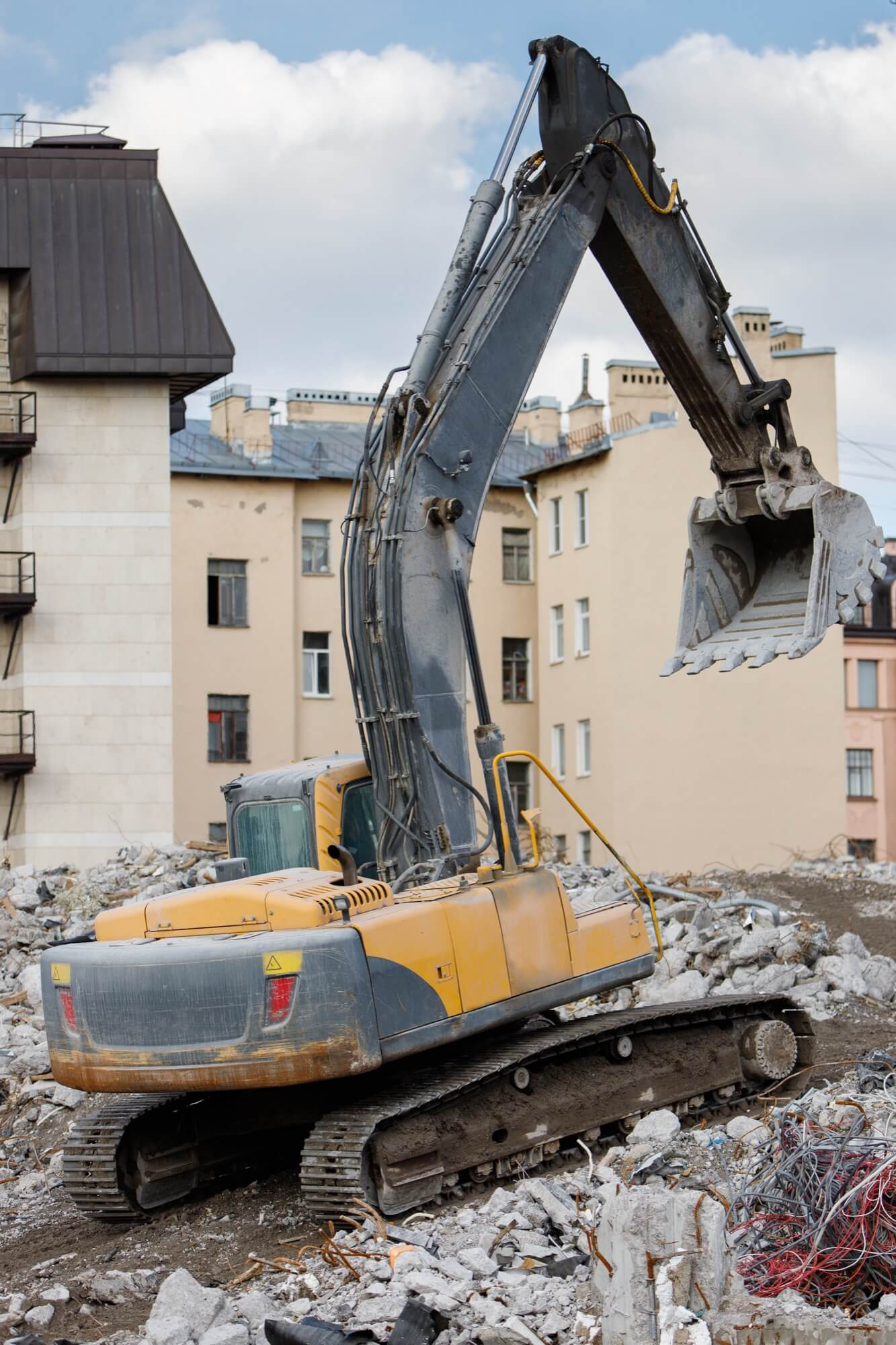 En grävmaskin på larvfötter positionerad på en hög av betong från en rivning i en tätbebyggd stadsmiljö.